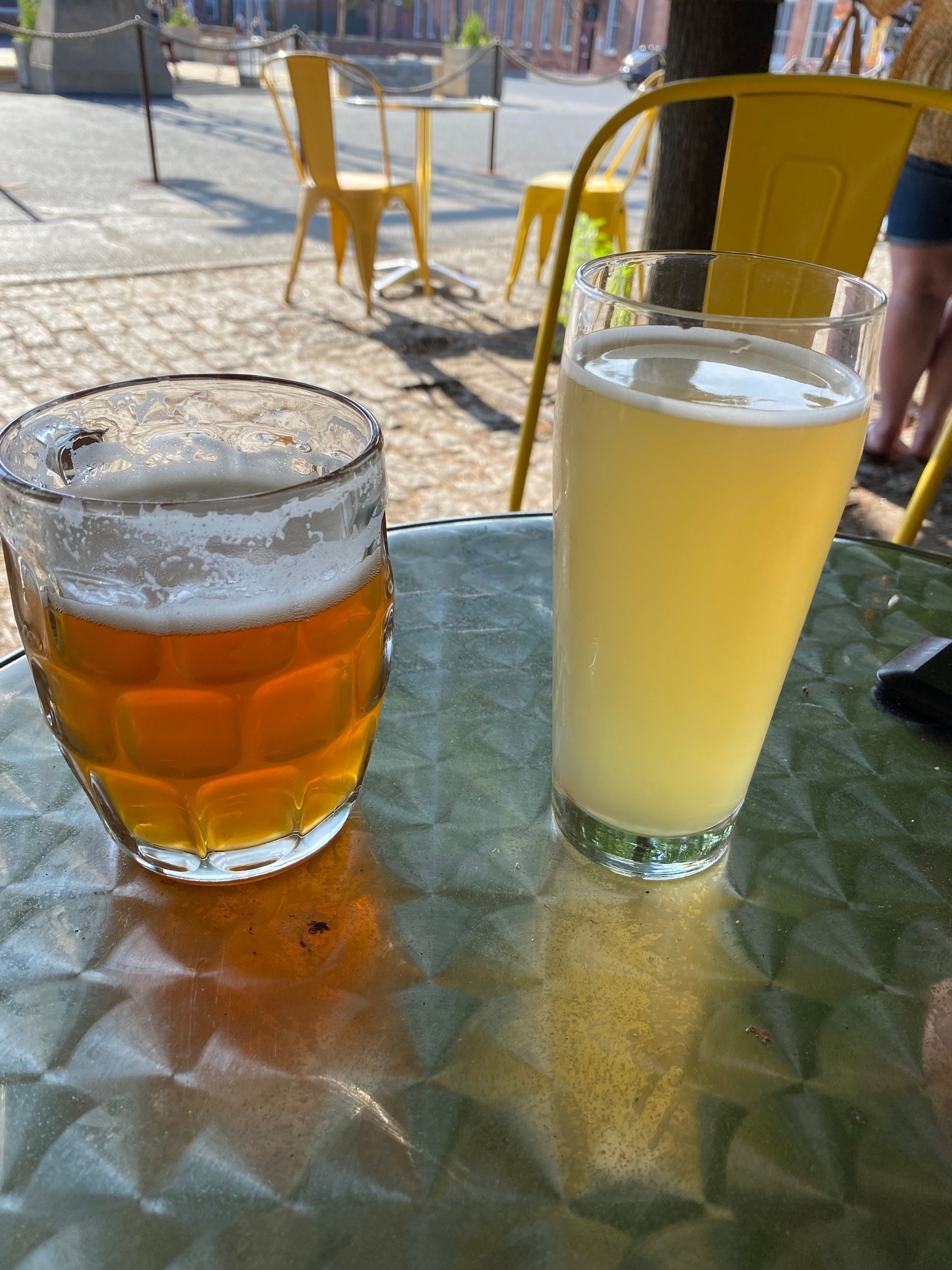 Two pints of beer on a metal table with yellow chairs in the background.