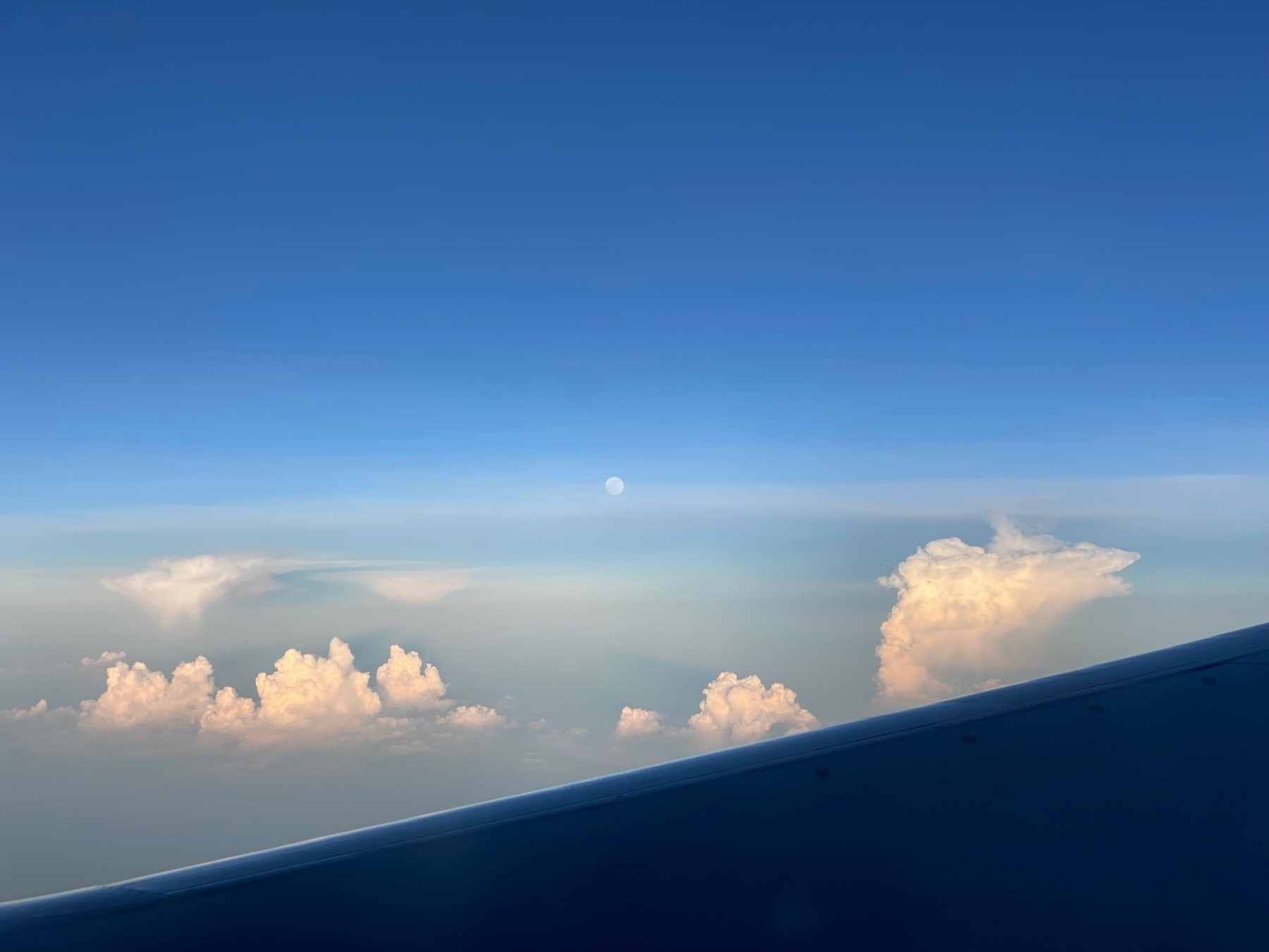 Clouds lit by the setting sun from the window of an airplane, with the nearly full moon just above the horizon.