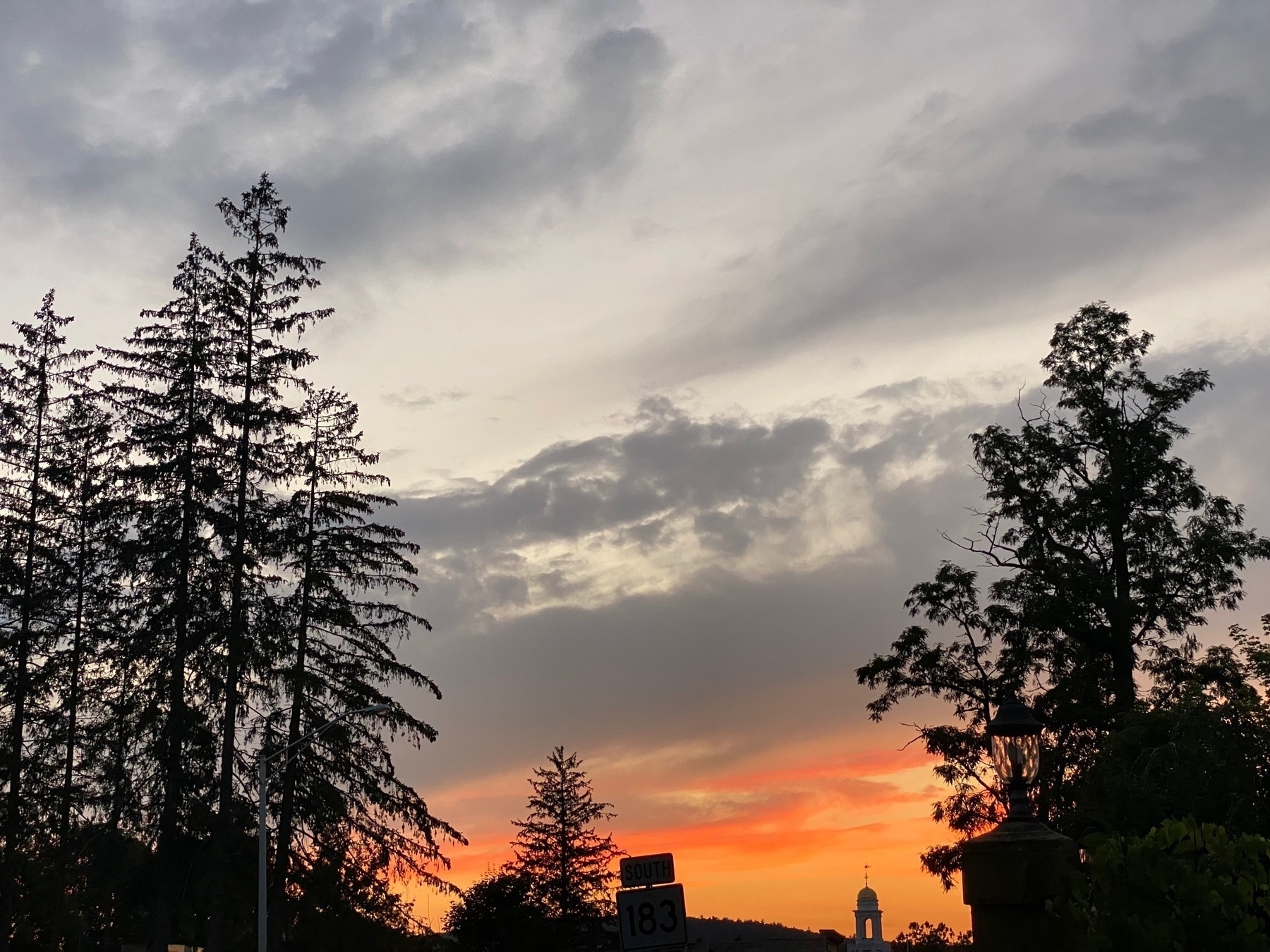 View of a sunset with grey clouds above and orange clouds below, silhouetted by trees on either side.