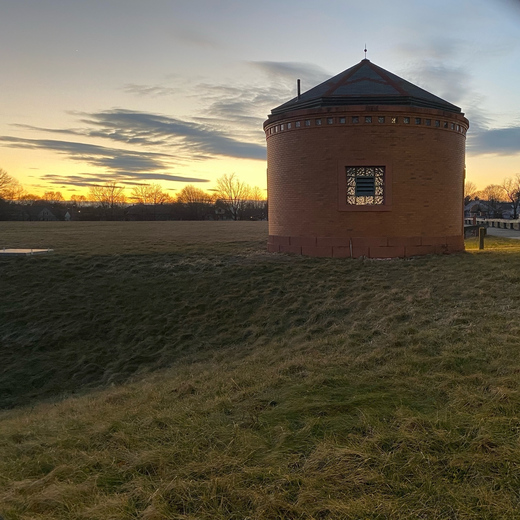 Vies of a small round brick building with an empty field and sunset behind it.