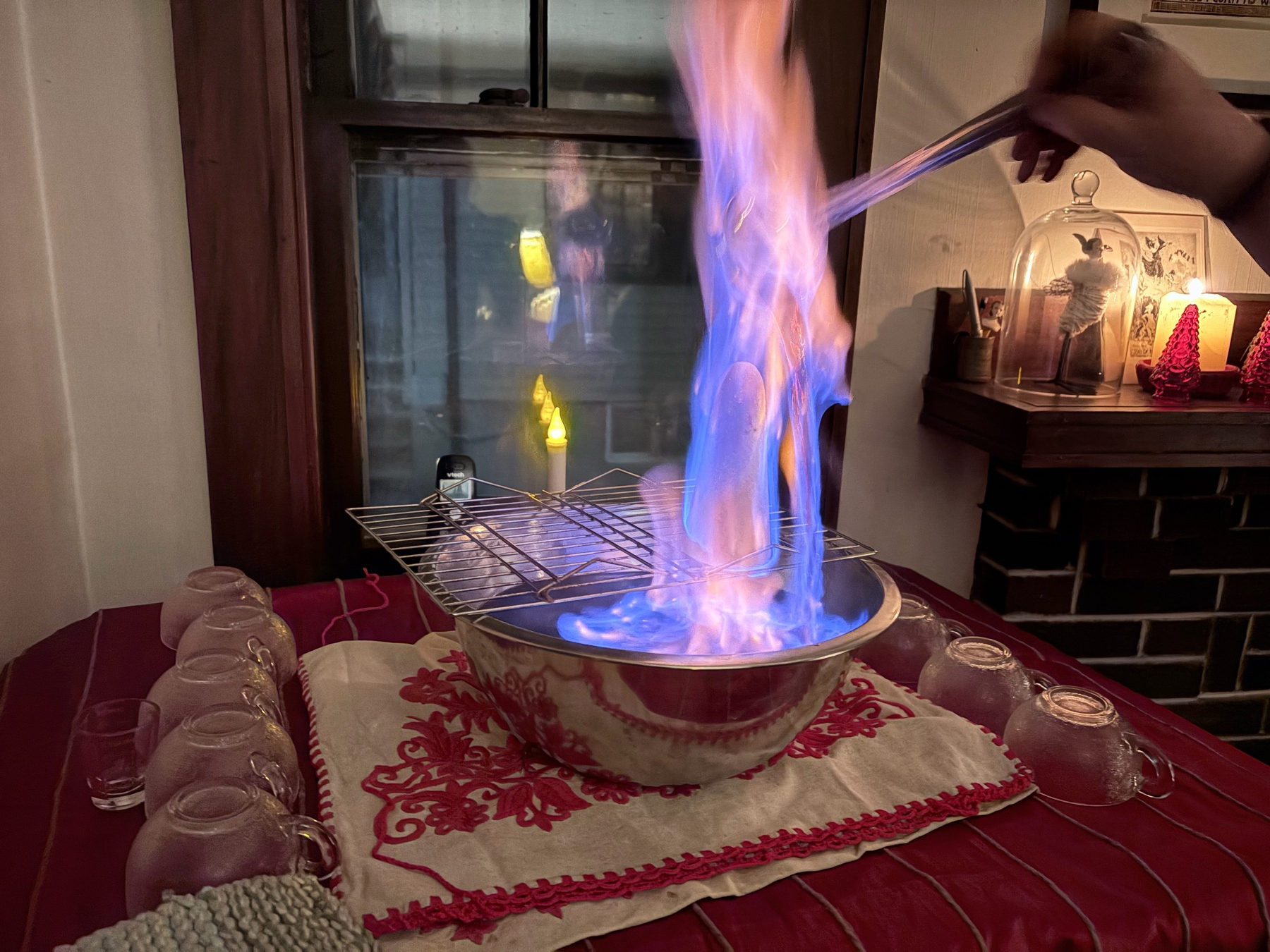 Flames rising from a metal bowl with a sugar cone on a wire rack on top, a hand holding a ladle on the right pouring liquid on the cone, a dark window in the background reflecting a small electric candle.